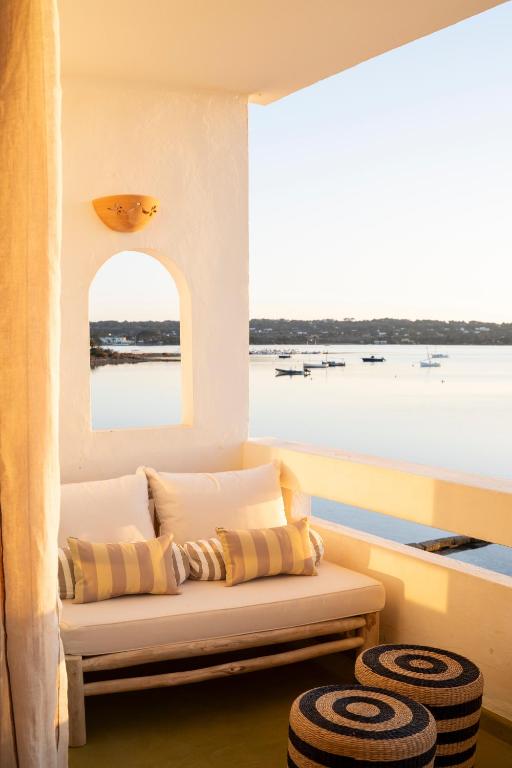 A cozy balcony in the style of boutique hotels in Formentera features a cushioned bench with striped pillows, overlooking calm waters dotted with boats under golden sunlight. Two round stools beside the bench complete this tranquil, inviting scene.