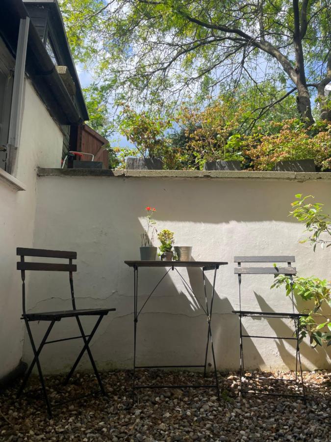 15 Best Boutique Hotels in London, UK 13 Two empty black metal chairs and a small table with potted plants sit on a gravel patio beside a white wall, evoking the charm of the best boutique hotels London has to offer, with green trees and sunlight in the background.