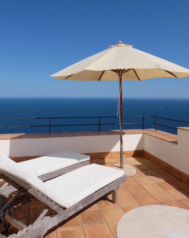 Two white cushioned lounge chairs and a large beige umbrella on a tiled terrace overlook the calm blue ocean—a scene reminiscent of boutique hotels in Costa Brava. Black railings border the terrace, with a small round table in the foreground.