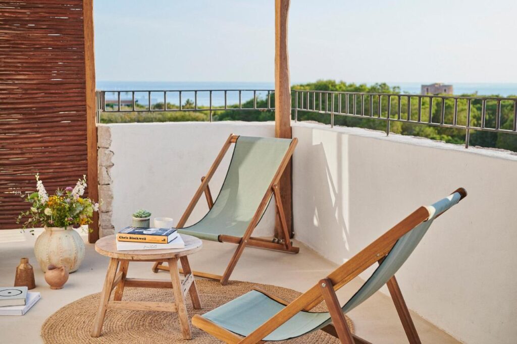 Two wooden deck chairs with light blue fabric sit on a balcony with a round mat, small table, books, and flowers—an inviting spot that captures the relaxed charm found at boutique hotels in Formentera, overlooking greenery and the distant sea.