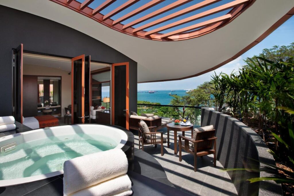13 Most Unique Hotels in Singapore for an Incredible Holiday 55 A luxurious balcony at one of the unique hotels in Singapore, featuring a round outdoor jacuzzi, lounge chairs, and a small dining table, overlooking the ocean and surrounded by greenery under a modern, partially covered roof.