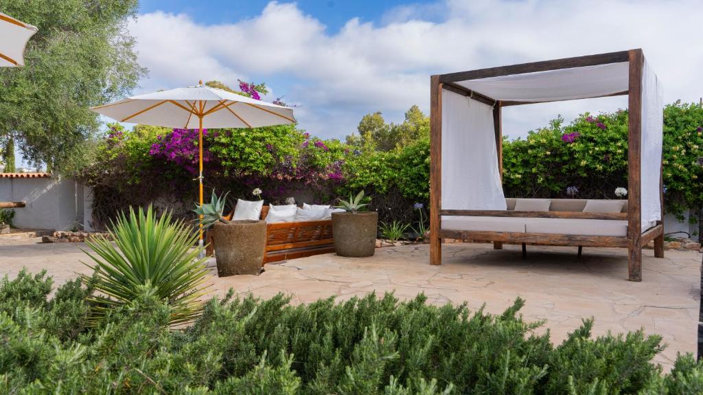 A sunny outdoor patio, inspired by boutique hotels in Formentera, features a wooden canopy bed with white curtains, a cushioned bench, large potted plants, an umbrella, and lush flowering greenery along the fence under a partly cloudy sky.