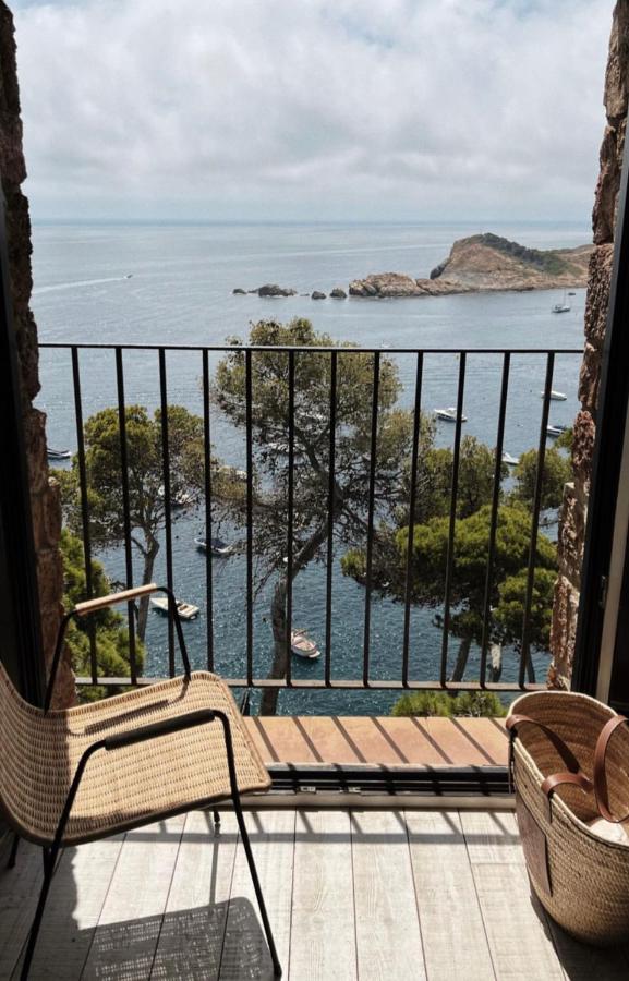 A balcony at one of the boutique hotels in Costa Brava features two wicker chairs overlooking the sea, boats, rocky islands, and lush green trees under a partly cloudy sky.