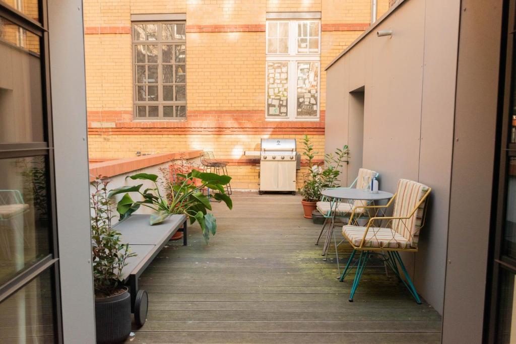 Boutique Hotels in Berlin Kreuzberg 25 A cozy outdoor patio, inspired by boutique hotels in Berlin Kreuzberg, features wooden flooring, potted plants, a bench, a small dining table with two chairs, and a barbecue grill against a brick wall bathed in natural light.