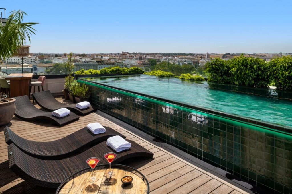 10 UNIQUE Hotels with Pool in Seville 5 A rooftop infinity pool with green tiles overlooks a cityscape, echoing the style of top hotels with pool in Seville. Black wicker lounge chairs and a snack table create the perfect spot to relax under the blue sky.