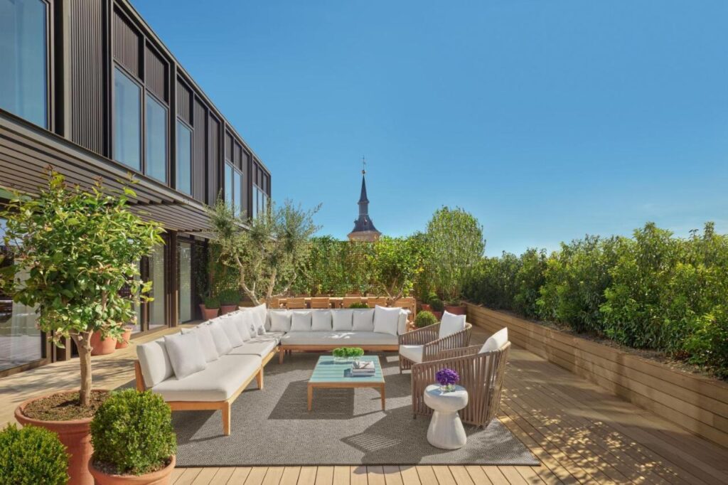 Where to Stay in Madrid: 14 Best Hotels 23 A spacious rooftop terrace with wooden decking, modern outdoor furniture, potted plants, and lush greenery—an inviting spot for those wondering where to stay in Madrid first time. A historic building with a pointed spire rises in the background under clear blue skies.
