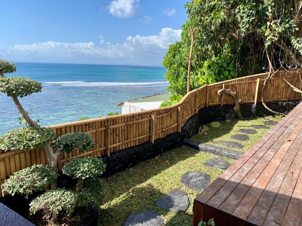 A wooden deck overlooks a lush garden with stepping stones, a bamboo fence, and stunning views of the turquoise ocean—perfect for relaxing at one of the best family villas in Bali.