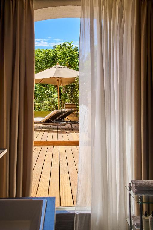 11 Unique Places to Stay in Provence 36 View through sheer curtains of a sunny wooden deck with two lounge chairs and a large umbrella, surrounded by greenery and blue sky—one of the unique places to stay in Provence.
