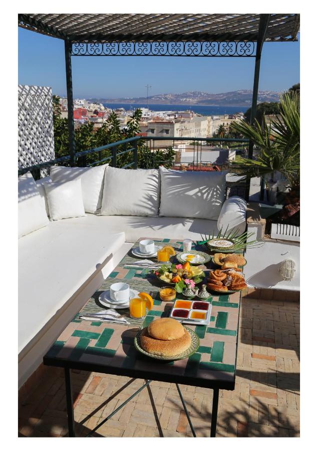 9 UNIQUE Riads in Tangier 13 A rooftop patio, reminiscent of the elegant riads in Tangier, features a white cushioned sofa and a table set for breakfast—pancakes, orange juice, bread, fruit, and pastries—overlooking the city and sparkling water beyond.