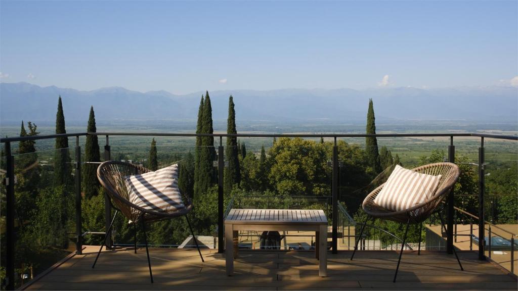 11 Unique Hotels in Georgia You Can't Miss! 37 Two wicker chairs with striped pillows and a small table sit on a balcony with glass railings, overlooking lush trees and distant mountainsāa serene retreat reminiscent of the views at unique hotels in Georgia.