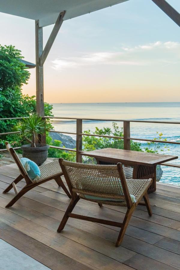 Two wooden lounge chairs and a table on a deck overlook the ocean at sunset, surrounded by greenery and a wooden railing—offering a peaceful, relaxing outdoor setting often found at the best family villas in Bali.