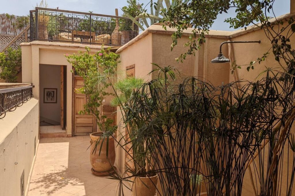 12 Charming Budget Riads in Marrakech 44 An outdoor shower with a metal showerhead is mounted on a tan wall, surrounded by lush potted plants. A small staircase leads to a door beneath the rooftop terrace, evoking the charm of budget riads in Marrakech.