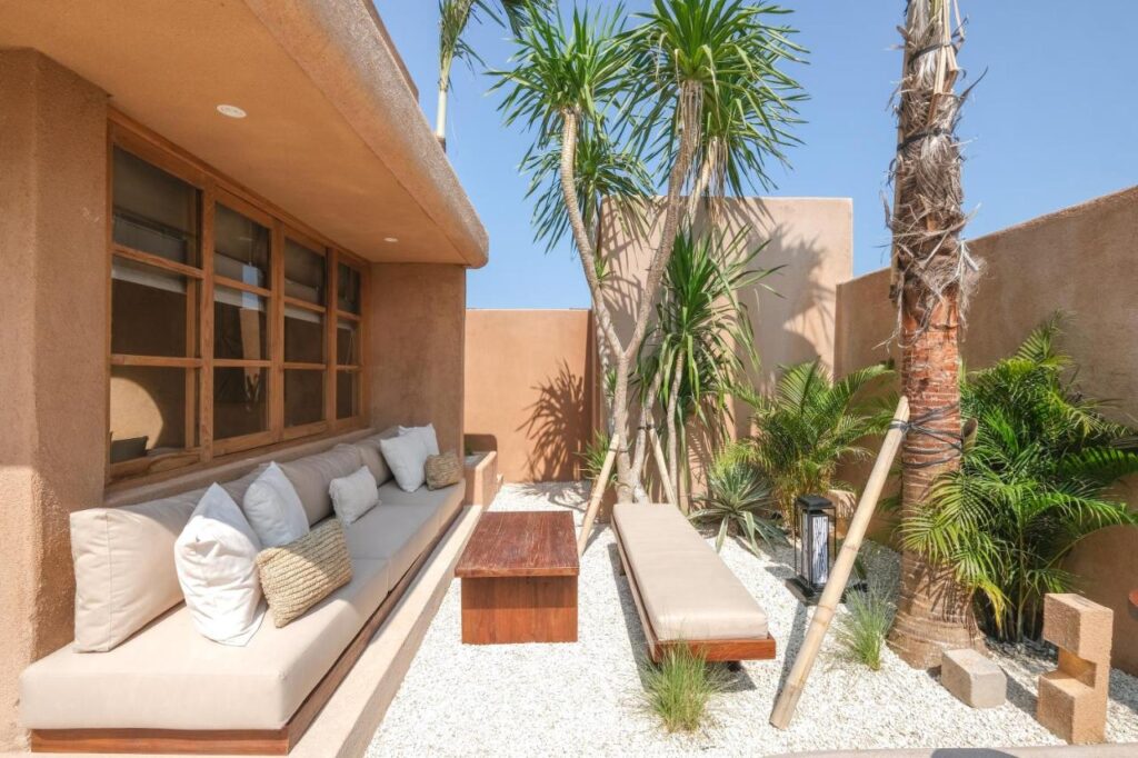 Sunny outdoor patio with beige cushioned seating, wooden table, benches, tall palm trees, and green plants on white gravel—just like the serene spaces found in the best family villas in Bali—surrounded by tan stucco walls under a clear blue sky.