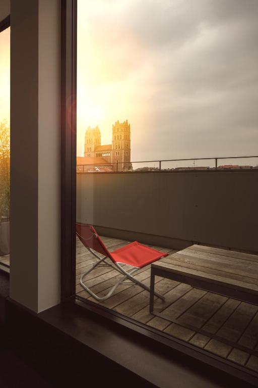 Authentic Boutique Hotels in Munich 5 A red lounge chair sits on a balcony with wooden flooring, overlooking a cityscape at sunset. In the distance, two tall church towers rise—a view often admired from boutique hotels in Munich.