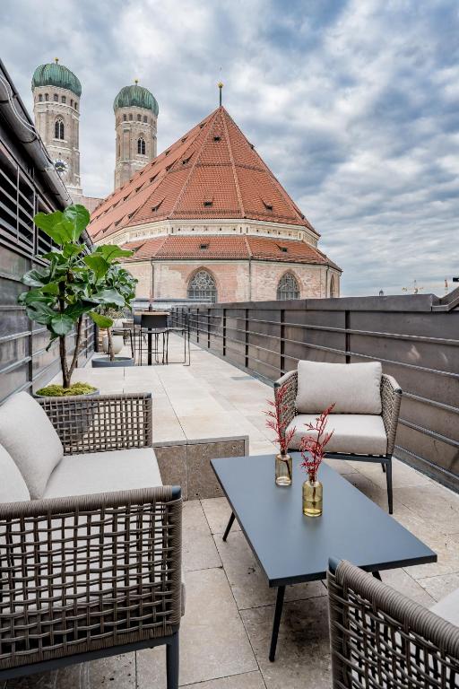 Authentic Boutique Hotels in Munich 22 A modern rooftop terrace at one of the boutique hotels in Munich features wicker chairs, small tables, and potted plants, overlooking a large historic church with a red-tiled roof and green domed towers under a cloudy sky.