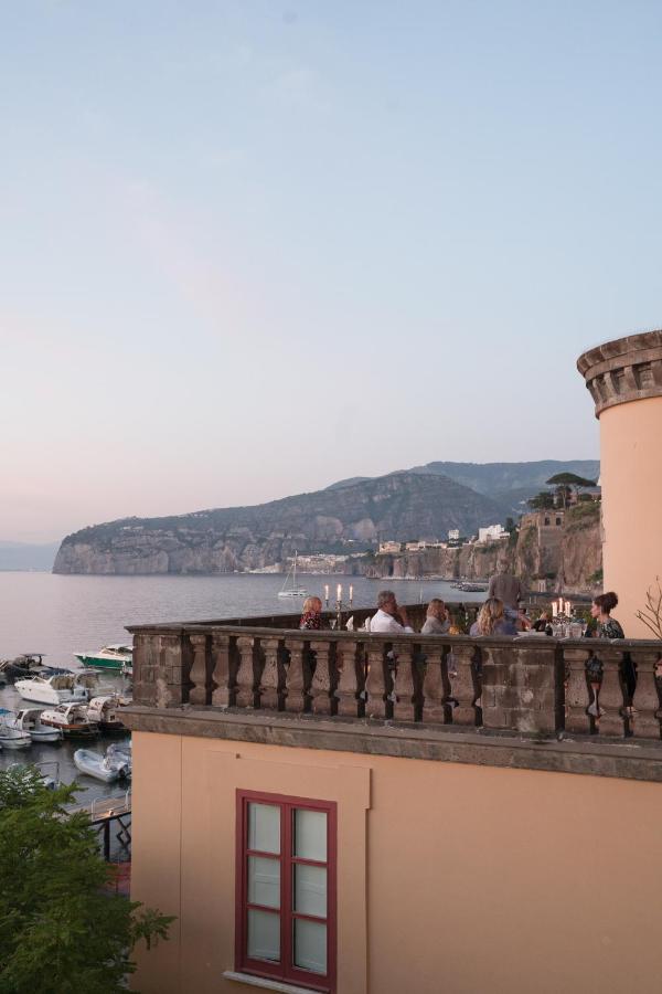 Boutique Hotels on the Amalfi Coast 5 A group of people sit on a terrace overlooking a marina and calm sea at sunset, with hills and boutique hotels on the Amalfi Coast visible in the background. The scene appears peaceful and scenic.