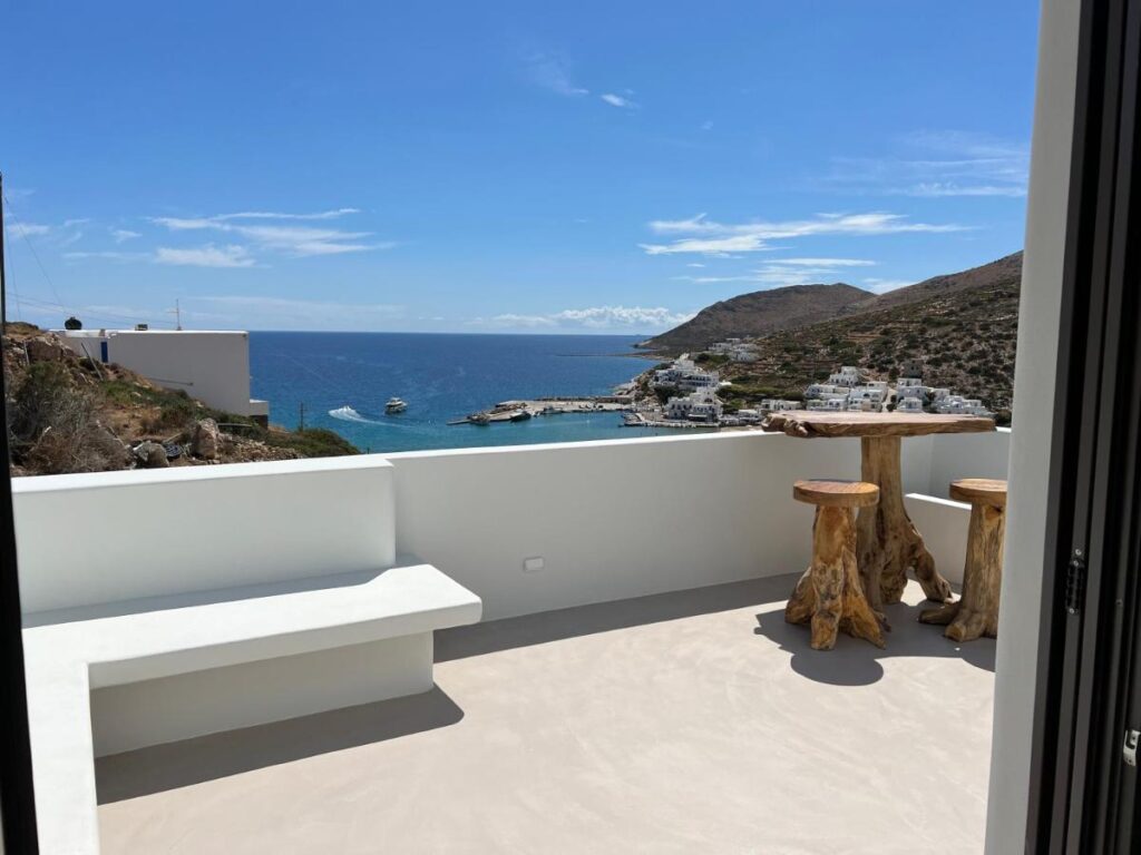 Authentic Hotels in Sikinos, Cyclades 3 A bright patio in one of the charming hotels in Sikinos features a white bench and rustic wooden stools, overlooking a coastal village, blue sea, and distant hills under a mostly clear sky.