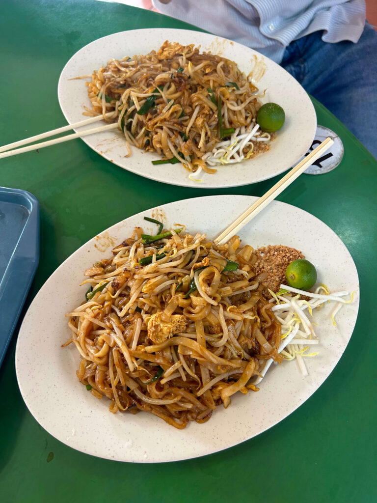 13 Most Unique Hotels in Singapore for an Incredible Holiday 60 Two plates of stir-fried noodles with bean sprouts, eggs, and vegetables are served on a green table—perfect for foodies exploring unique hotels in Singapore—each with chopsticks, a halved lime, and crushed peanuts on the side.