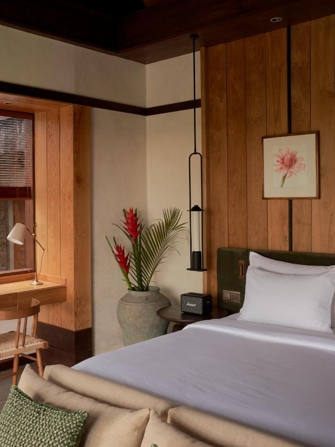 13 Most UNIQUE Places to Stay in Ubud 44 A cozy bedroom with a neatly made bed, a framed flower picture on a wooden wall, and potted plants evokes the charm of unique places to stay in Ubud. A wooden desk by the window offers a tranquil spot to relax or work.