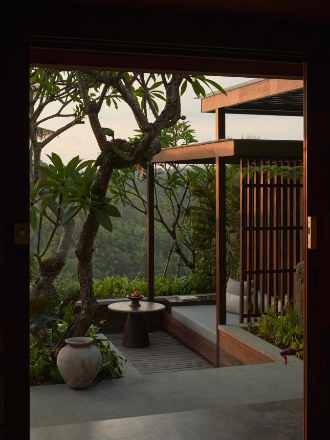 13 Most UNIQUE Places to Stay in Ubud 42 A serene outdoor patio with a wooden pergola, cushioned seating, a small round table, potted plants, and tropical trees—one of the unique places to stay in Ubud—overlooking lush greenery in the background at sunset.