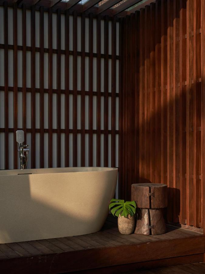 13 Most UNIQUE Places to Stay in Ubud 43 A modern bathroom with a freestanding bathtub, wooden stool, and a small green potted plant, set against vertical wooden slats with sunlight casting shadows—perfect for those seeking unique places to stay in Ubud.