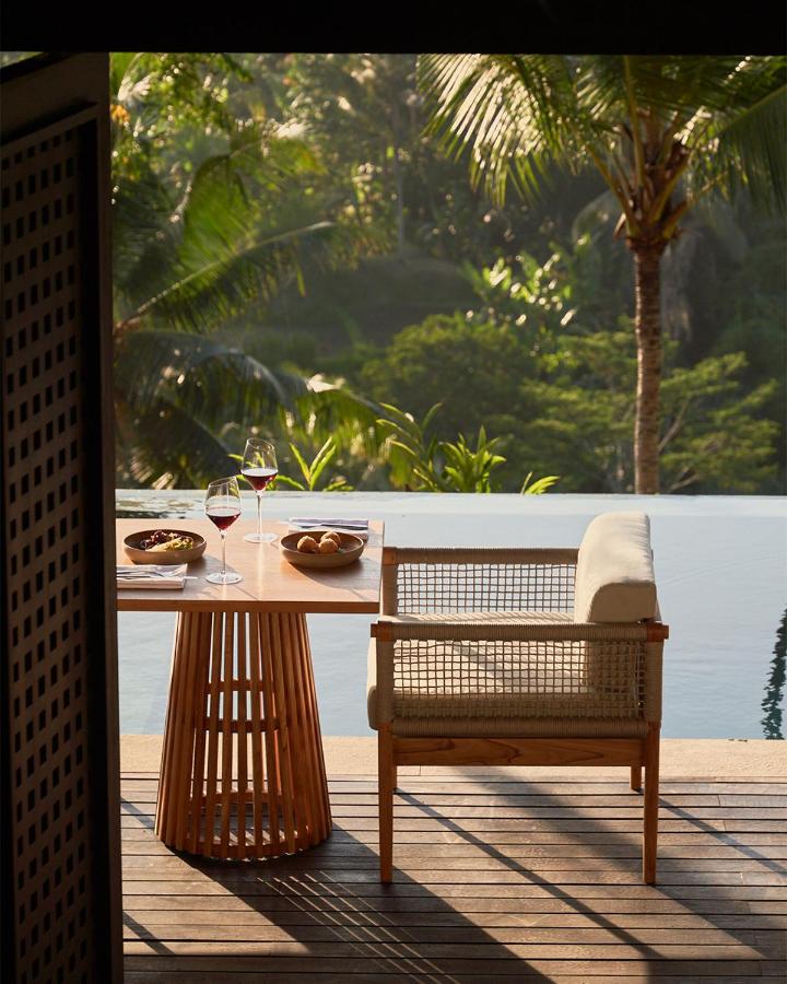 13 Most UNIQUE Places to Stay in Ubud 45 A cozy outdoor dining setup with a wooden table and chair by a pool, featuring two glasses of red wine, bowls of snacks, and lush tropical greenery—perfect for those seeking unique places to stay in Ubud.