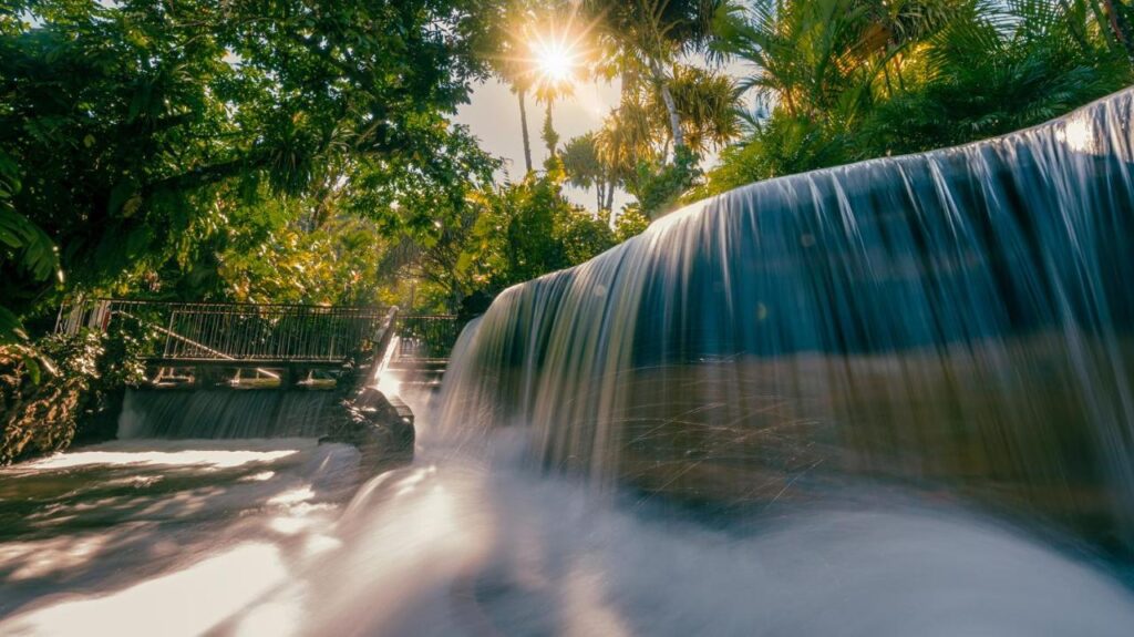 Unique Hotels in Costa Rica: Top Stays 3 A waterfall flows smoothly over rocks, surrounded by lush green trees and plants. Sunlight shines through the foliage, creating a serene atmosphere—much like the views from unique hotels in Costa Rica, with a bridge visible in the background.