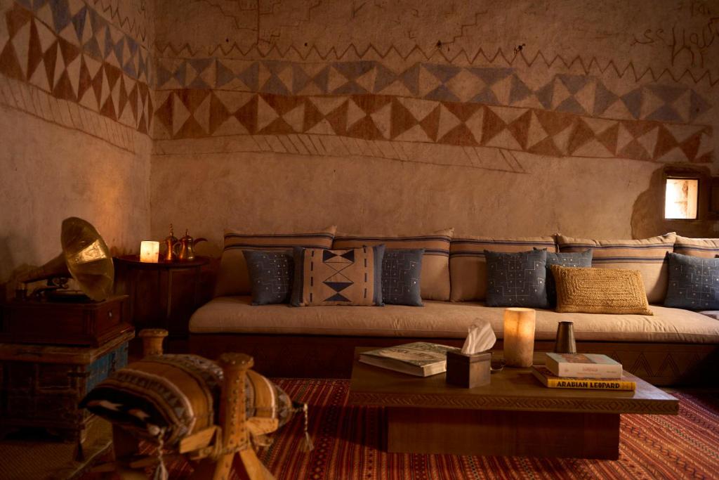 9 Best Hotels in Saudi Arabia: Unique Stays Across the Country 20 A cozy living room, reminiscent of the Best Hotels in Saudi Arabia, with a long cushioned sofa adorned with patterned blue and beige pillows, a wooden coffee table topped with books and candles, traditional decor, and geometric murals accenting the walls.