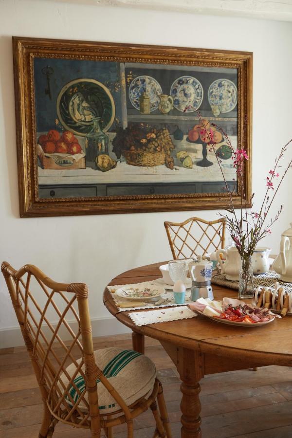25 Weekend Trips from Madrid Max. 2h Away 37 A round wooden dining table set for tea with pastries, a flower vase, and teapots sits before a wall adorned by a large still life painting—an inviting scene reminiscent of cozy weekend trips from Madrid. Two wicker chairs complete the setting.