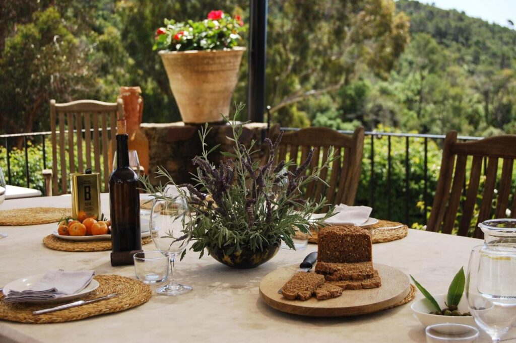 A rustic outdoor dining table set with plates, glasses, a bottle of wine, a loaf of bread, fresh fruit, and lavender evokes the charming ambience found at boutique hotels in Costa Brava, surrounded by wooden chairs and lush greenery.