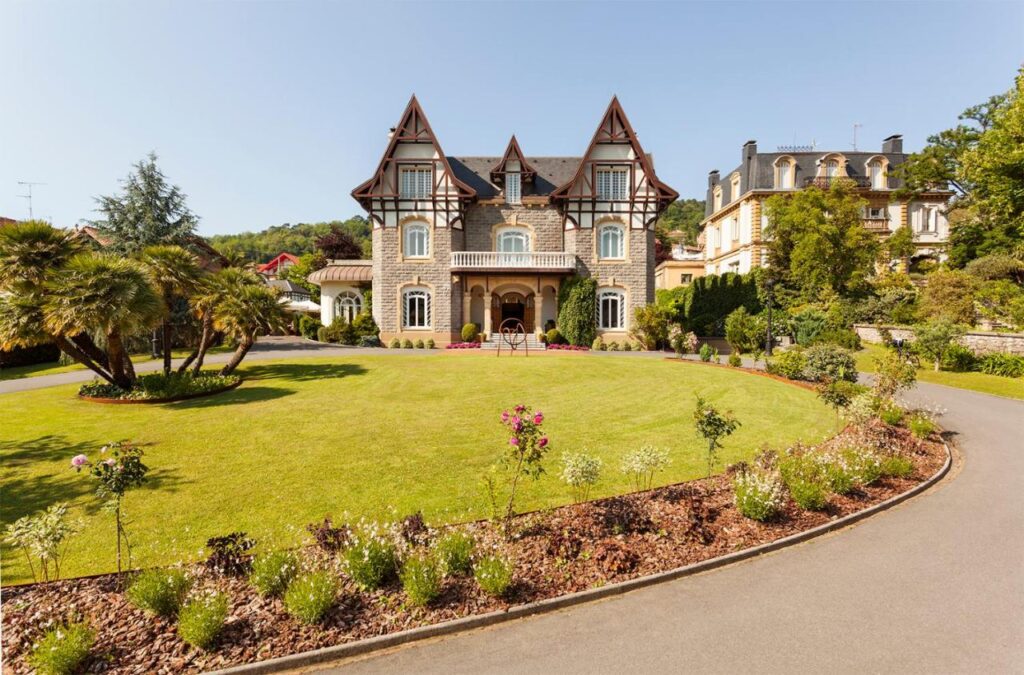 Best Boutique Hotels in San Sebastian 14 A grand, three-story stone mansion with pointed roofs and tall windows, reminiscent of the elegant boutique hotels in San Sebastian, set behind a circular driveway and surrounded by manicured lawns, flower beds, and palm trees on a sunny day.