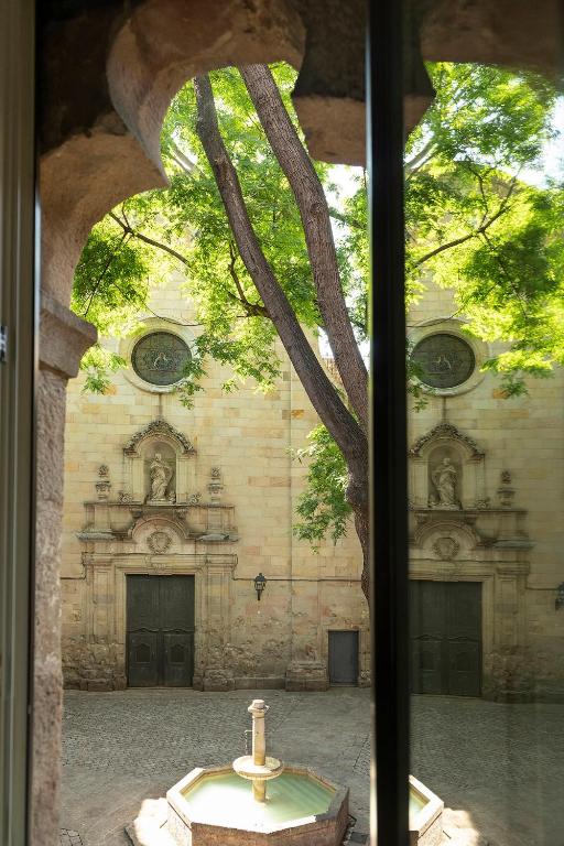 13 Unique Hotels in Barcelona, Spain 44 A stone courtyard with a central fountain is seen through a window. Like many Barcelona hotels with rooftop pool access, an old building with arched doors and circular windows stands nearby, its tree casting shadows on the sunlit stone facade.