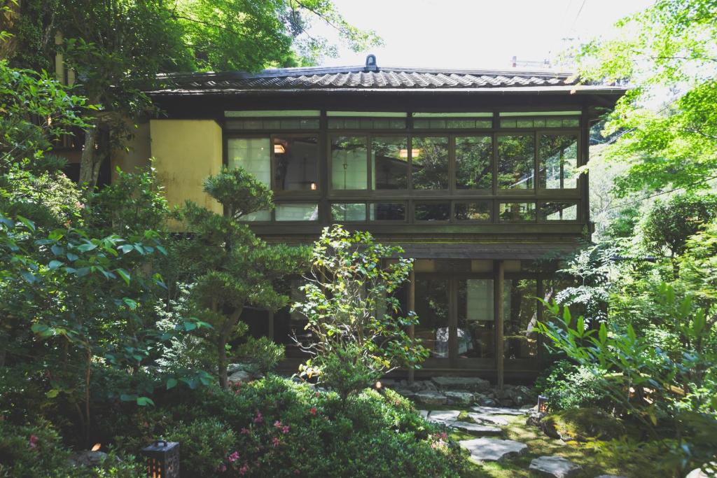 8 BEST Ryokans in Osaka 31 A traditional Japanese house with large glass windows is surrounded by lush green trees and a garden path, evoking the peaceful and serene atmosphere found at ryokans in Osaka under natural sunlight.