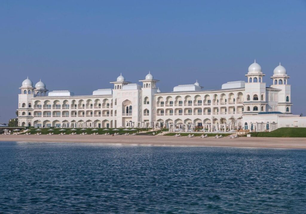 Where to Stay in Doha, Qatar: Transit, City & Beach Hotels 7 A large, white palace-like building with domed towers and arches stands on a sandy beach by calm blue water under a clear sky—an ideal spot if you're wondering where to stay in Doha, with rows of lounge chairs and umbrellas lining the beachfront.