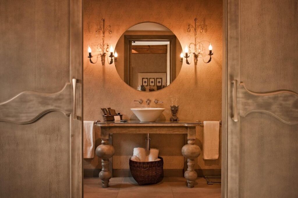 25 Weekend Trips from Madrid Max. 2h Away 87 Warmly lit bathroom with beige walls, a round mirror above a vessel sink, ornate sconces on either side, a wooden vanity with towels—perfect for relaxing after weekend trips from Madrid—plus a wicker basket beneath and double doors framing the view.