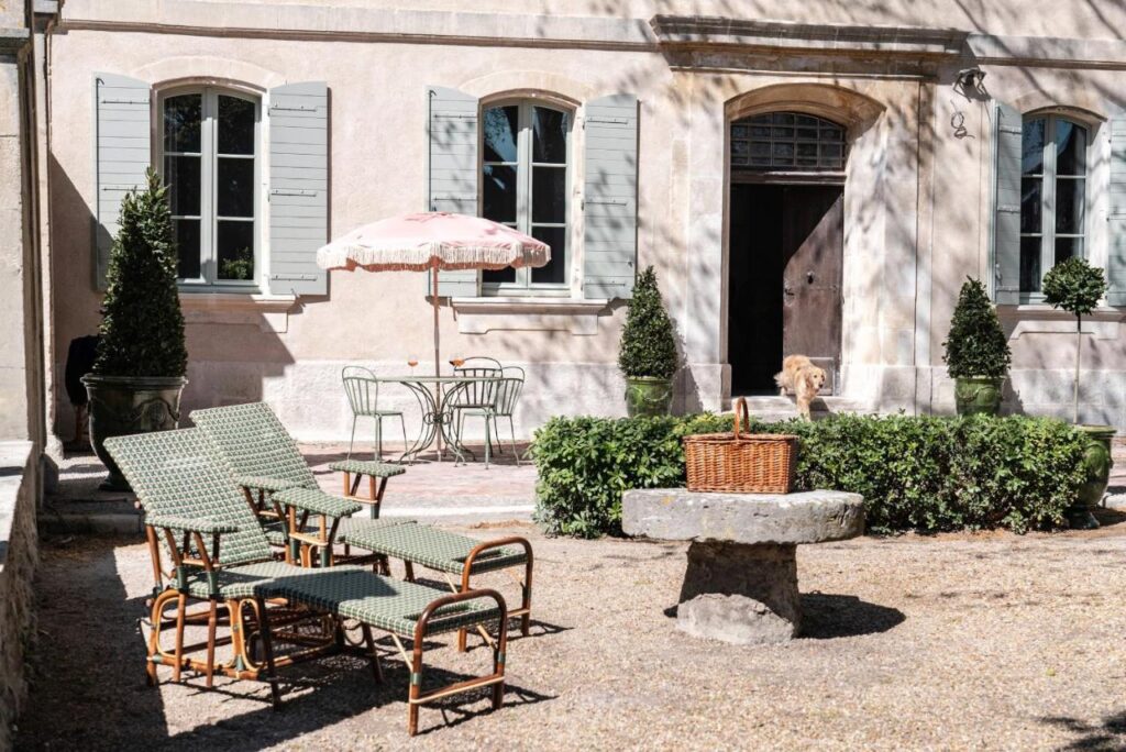 11 Unique Places to Stay in Provence 3 A sunny courtyard with green lounge chairs, a round metal table and chairs under a pink umbrella, trimmed bushes, and a light-colored building in the background—a charming scene among unique places to stay in Provence. A dog stands near a doorway.