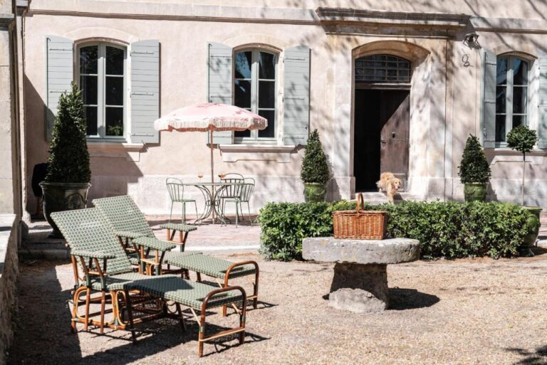 A sunny courtyard with green lounge chairs, a round metal table and chairs under a pink umbrella, trimmed bushes, and a light-colored building in the background—a charming scene among unique places to stay in Provence. A dog stands near a doorway.
