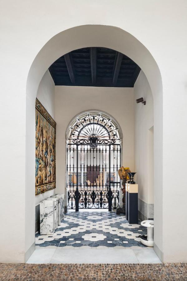 10 UNIQUE Hotels with Pool in Seville 34 A view through an arched doorway into a well-lit hall with patterned tile flooring—reminiscent of elegant hotels with pool in Seville—featuring a tapestry, marble tables, sculptures, and an ornate iron gate before a sunlit window.