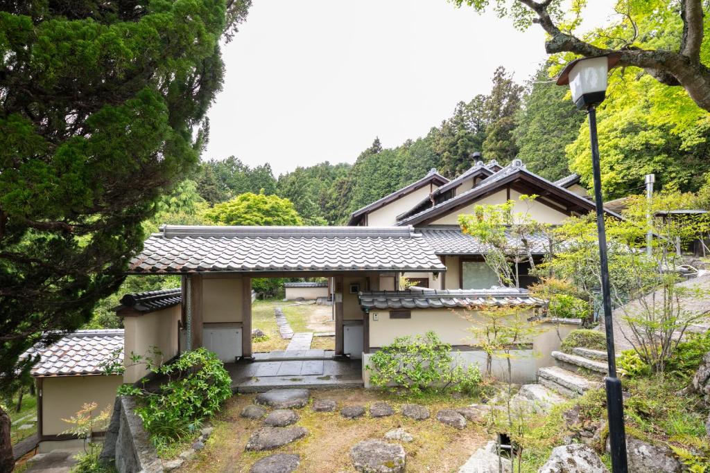 8 BEST Ryokans in Osaka 26 Traditional Japanese house with a tiled roof, like the charming ryokans in Osaka, surrounded by lush greenery and trees. A stone path leads to the entrance gate, and steps with a lamppost rise to the right in this peaceful setting.