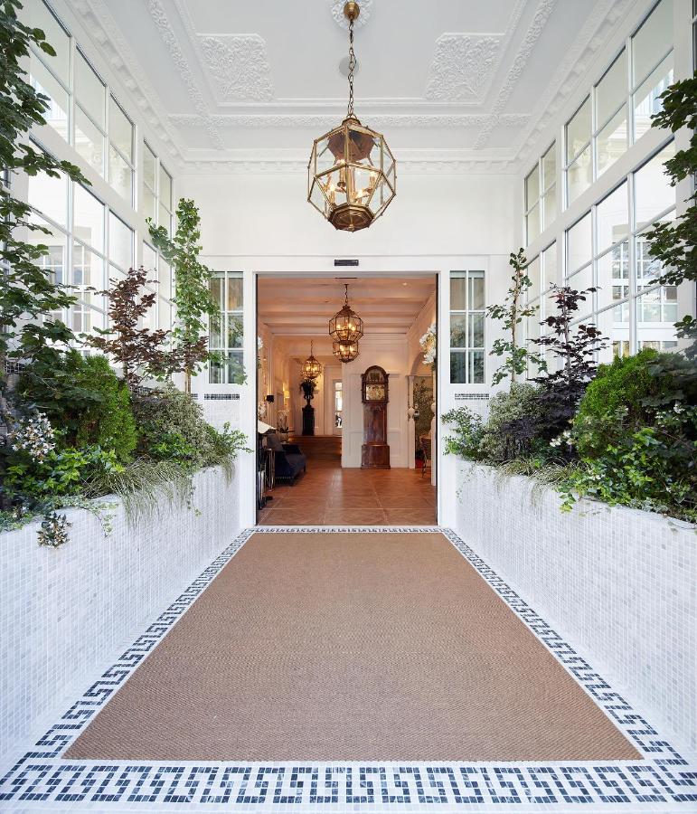 Best Boutique Hotels in San Sebastian 18 Elegant entryway with white walls, large windows, and greenery in raised planters—much like the style found in boutique hotels in San Sebastian. Ornate hanging lights and a detailed ceiling open into a warmly lit space with wooden floors.
