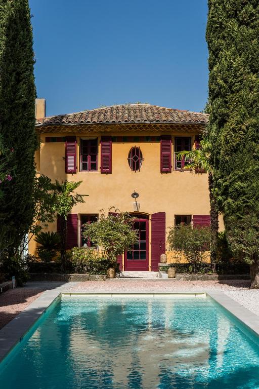 11 Unique Places to Stay in Provence 34 A yellow two-story house with red shutters and a tiled roof stands behind a rectangular swimming pool, framed by tall green trees and shrubs under a clear blue sky—one of the unique places to stay in Provence.