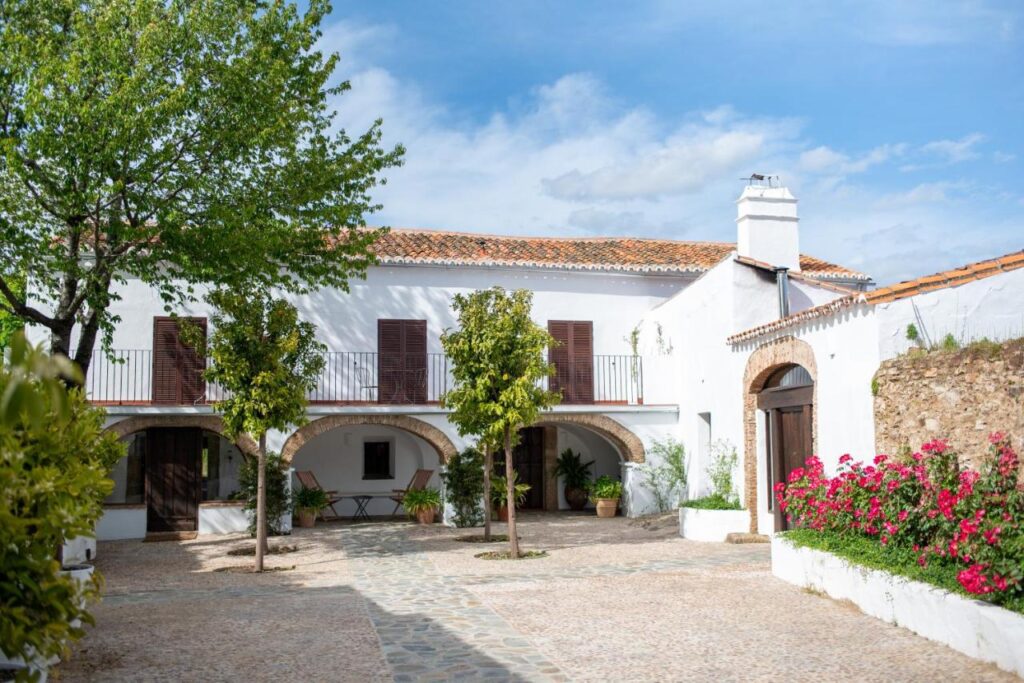 25 Weekend Trips from Madrid Max. 2h Away 12 A white, two-story Mediterranean-style house with wooden shutters, arched doorways, and a tiled roof—perfect for weekend trips from Madrid—surrounded by trees, pink flowers, and a cobblestone courtyard under a blue sky.