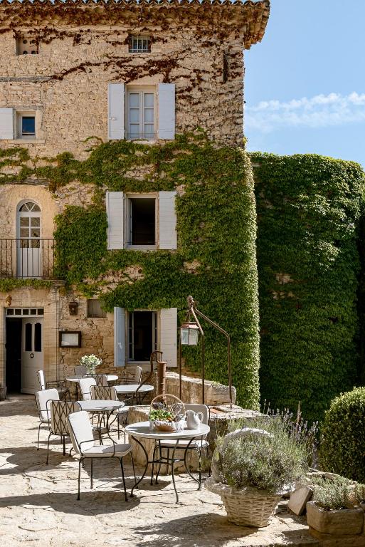 11 Unique Places to Stay in Provence 32 A charming stone building with white shutters and ivy climbing its walls, this retreat is one of the unique places to stay in Provence, featuring an outdoor patio with white chairs, round tables, potted plants, and a clear blue sky above.