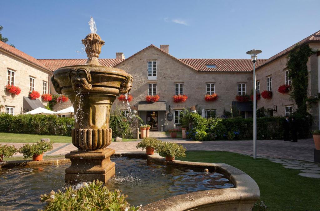 A stone courtyard features a large ornate fountain in the foreground, with a historic building—reminiscent of boutique hotels in Galicia—behind it. Red flowers hang below the windows, and greenery surrounds the scene under a clear blue sky.