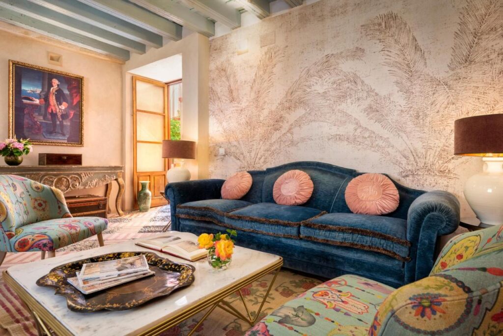 10 UNIQUE Hotels with Pool in Seville 33 Cozy living room with a blue velvet sofa, pink round pillows, floral armchairs, and a marble coffee table. Inspired by hotels with pool in Seville, the space features sunlit palm leaf wall murals, stylish wall art, and elegant touches throughout.