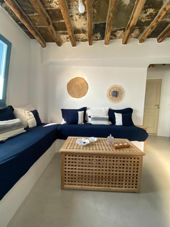 Authentic Hotels in Sikinos, Cyclades 7 A cozy living room, reminiscent of boutique hotels in Sikinos, features navy blue sofas, white and blue pillows, a wooden coffee table, rustic wooden ceiling beams, and round woven wall decor. Sunlight streams in from a window on the left.
