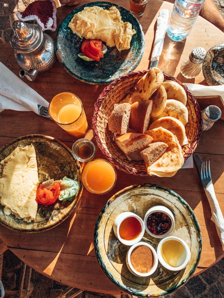 12 Charming Budget Riads in Marrakech 4 A table set for breakfast at one of the budget riads in Marrakech features plates of omelets, sliced tomatoes, assorted bread in a basket, spreads, orange juice, a metal teapot, bottled water, and napkins bathed in bright sunlight.