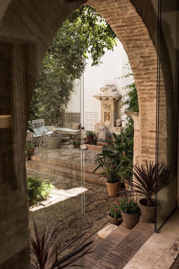 Best of Cordoba One Day Itinerary 3 A view through a stone archway reveals a serene courtyard in Cordoba—perfect for a one day visit—with potted plants, cobblestone ground, rustic seating, and an old stone fountain surrounded by greenery.