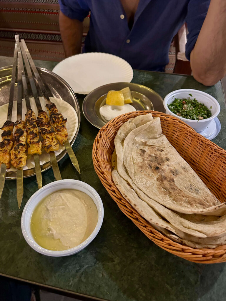 Grilled kebabs on skewers, flatbreads in a basket, hummus, parsley salad, and lemon wedges with sauce set on a green table; a seated person in a blue shirt is partially visible—a scene to enjoy while exploring where to stay in Doha.
