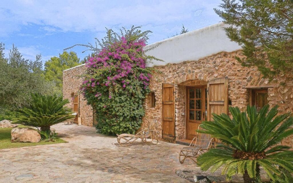 10 AUTHENTIC Finca Hotels in Ibiza 41 A rustic stone house with wooden doors and windows, reminiscent of finca hotels in Ibiza, is surrounded by lush greenery and flowering vines. Two wicker lounge chairs rest on a stone patio beneath a partly cloudy sky.